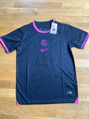 Svart PSG fotbollströja Nike - Snygg svart PSG fotbollströja från Nike med rosa detaljer vid krage och ärmslut. Tryck med klubbmärke och Nike-logga framtill, samt D. Doué och nummer 14 på ryggen i lila. Tillverkad i lätt och ventilerande material, perfekt för träning eller match. Print fel där bak