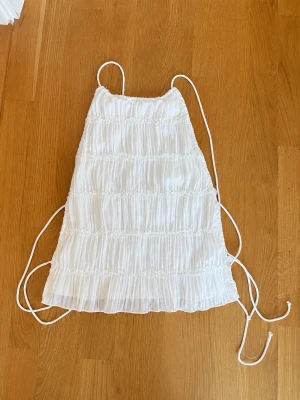 Vit linnetopp från Gina Tricot - Säljer en söt vit linnetopp från Gina Tricot med tunna axelband och smockade detaljer. Toppen har en luftig och flowy passform med knytband i sidorna, perfekt för varma dagar. Materialet känns lätt och somrigt.