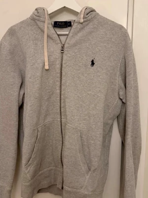 Grå zip hoodie från Polo Ralph Lauren - Snygg grå hoodie med dragkedja från Polo Ralph Lauren. Klassisk design med huva och vita snören, två fickor framtill och den ikoniska loggan broderad på bröstet. Perfekt för en chill och avslappnad stil. Lite rost på zippers annars lungt