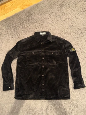 Svart manchester overshirt Stone Island - Säljer en svart overshirt i manchestertyg från Stone Island. Skjortan har två bröstfickor med knappar, klassisk krage och ikonisk patch på vänster ärm. Perfekt för lager-på-lager och ger en clean-look.