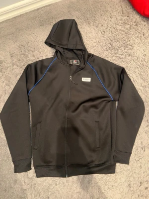 Svart hoodie från McKenzie med blå detaljer - Säljer en svart hoodie från McKenzie med dragkedja framtill och blå detaljer längs axlarna. Hoodien har två fickor, huva och en liten McKenzie-logga på bröstet. Materialet är mjukt och sportigt, perfekt för en avslappnad stil.