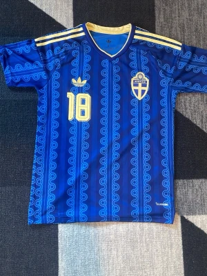 Blå Sverige fotbollströja Adidas - Säljer en blå fotbollströja från Adidas med Sveriges landslagsemblem och nummer 18 i gult. Tröjan har gula detaljer på axlarna och ett unikt mönster över hela tyget. Perfekt för dig som vill stötta Sverige på match eller träning. Storlek är M