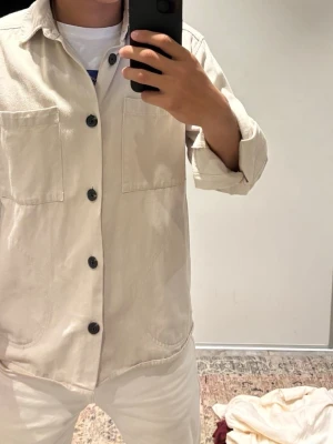 Beige overshirt från Isolid - Stilren beige overshirt från Isolid med två stora bröstfickor och svarta knappar. Overshirten har klassisk krage och raka ärmar med knapp vid ärmslut. Tillverkad i ett mjukt, vävt material som passar perfekt för lager på lager. Knappt använd. Storlek S men funkar på M. Väldigt snygg nu till hösten!!!