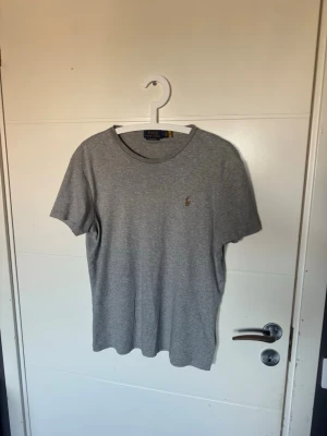 Grå t-shirt från Polo Ralph Lauren - Klassisk grå t-shirt från Polo Ralph Lauren med rund hals och korta ärmar. Liten broderad logga på bröstet i kontrastfärg. Tillverkad i mjuk bomull för en bekväm känsla och enkel stil.