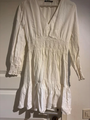 Vit kort klänning med v-ringning - Söt vit kort klänning med v-ringad hals, smock i midjan och långa ärmar med smockade muddar. Kjolen har volangkant och klänningen är tillverkad i ett lätt tyg som passar perfekt till varmare dagar.