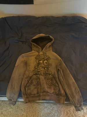 Cactus Jack X Takashi Murakami Hollow Hoodie - Cactus Jack hoodie med huva och stora grafiska tryck fram och bak. Framsidan har ett konstnärligt monster-motiv och baksidan visar flera figurer med texten 'HOLLOW'. Ärmen har en svart flammdesign. Hoodien har ribbade muddar och avslappnad passform.