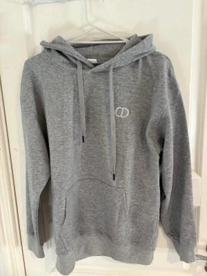 Grå hoodie med broderad logga - Snygg grå hoodie med broderad vit logga på bröstet. Hoodien har justerbar dragsko i huvan, stor känguruficka framtill och ribbade muddar vid ärmar och nederkant. Tillverkad i mjukt sweatshirtmaterial som är perfekt för chill dagar.