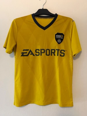 Gul FIFA 17 EA Sports tröja - Cool gul FIFA 17 Ultimate Team tröja med svart EA Sports-logga på bröstet och stort nummer 17 på ryggen. T-shirten har korta ärmar, svart V-ringad krage och är tillverkad i ett lätt och ventilerande material, perfekt för sport eller gaming.