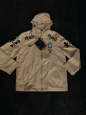 Vit Nike Brasilien jacka med GORE text  - Nike Brasilien Gore-Tex jacka 🇧🇷⚽️  Säljer en riktigt snygg Nike Brasilien-jacka i beige. Jackan har Brasilien/CBF-loggan på bröstet tillsammans med Nike-loggan och sponsordetaljer från Itaú, Vivo och Guaraná. Den har även Gore-Tex och är perfekt för både vardag och fotboll.  Jackan har huva, dragkedja hela vägen fram och en stilren beige färg som passar till det mesta. Väldigt snygg och ovanlig modell 🇧🇷  📏 Storlek: M 🎨 Färg: Beige 🧥 Märke: Nike 🇧🇷 Brasilien/CBF ☔ Gore-Tex ✨ Skick: Ny med prislapp 