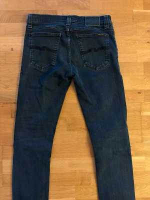 Nudie jeans - Snygga mörkblå jeans med raka ben och två bakfickor med vågformad söm. Jeansen har bälteshällor och klassisk femficksdesign. Materialet är denim i bomull och passformen är normal. Storlek 29:34