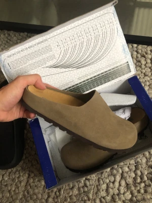 Beige mockatofflor med sula - Säljer ett par stilrena beige tofflor i mocka med sluten tå och öppen häl. Tofflorna har en robust sula som passar perfekt för både inomhus och snabba ärenden utomhus. Klassisk och enkel design som funkar till det mesta.
