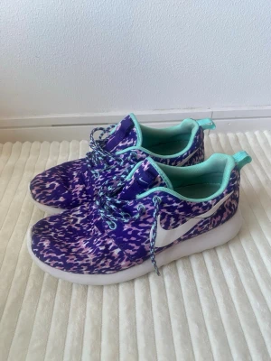 Lila mönstrade sneakers med turkosa detaljer - Säljer ett par sneakers med lila, vita och rosa mönster samt turkosa detaljer på insidan och snörningen. Skorna har en vit sula och en stor vit swoosh på sidan. Perfekta för dig som vill sticka ut med färg och mönster.