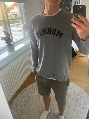 Grå stickad tröja från Morris - Snygg grå stickad tröja från Morris med svart broderad logga på bröstet. Tröjan har rund halsringning och ribbade muddar vid ärmslut och nederkant. Perfekt för en avslappnad och stilren look.
