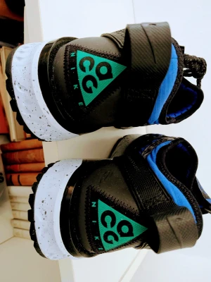 Nike ACG blå och svarta sneakers - Snygga Nike ACG sneakers i blått och svart med unikt snörningssystem och specklad vit sula. Skorna har ett sportigt utseende med robust yttersula och detaljerade svarta band över ovandelen. Perfekta för dig som gillar streetwear och outdoor-vibe.