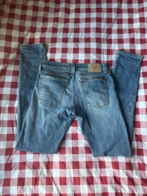 Nudie jeans - Säljer ett par ljusblå skinny jeans från Nudie med klassisk femficksdesign och läderpatch bak i midjan. Jeansen har smal passform och är tillverkade i mjukt denimtyg. Perfekta för en avslappnad och trendig look.
