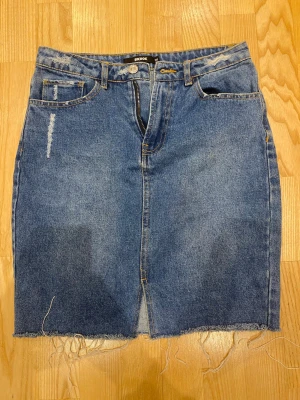 Blå jeanskjol med rå kant från Bik Bok - Säljer en blå jeanskjol från Bik Bok med rå, fransig kant och slits framtill. Kjolen har klassiska fem fickor, bälteshällor och slitna detaljer för en trendig look. Perfekt till sommarens outfits!