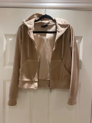 Beige velour hoodie med dragkedja - Snygg beige hoodie i mjuk velour med dragkedja framtill och två stora fickor. Har huva med snören och ribbade muddar vid ärmar och nederkant. Perfekt för en chill och avslappnad stil.