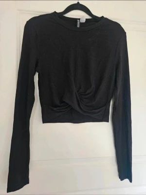 Svart croppad topp från H&M Divided - Svart croppad topp från H&M Divided  Säljer en svart långärmad croppad topp från H&M Divided. Toppen har en snygg twistad detalj framtill som ger en cool look. Mjuk och stretchig i materialet, perfekt att styla med höga jeans eller kjol.  Kan även hämtas I Partille  E1