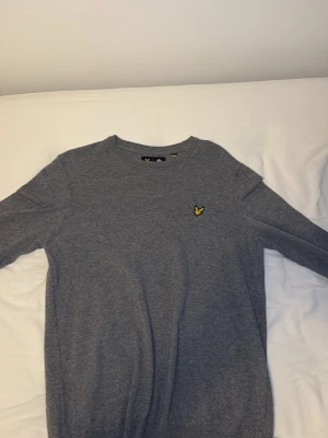 Grå stickad tröja från Lyle & Scott - Snygg grå stickad tröja från Lyle & Scott med rund halsringning och den klassiska gula loggan broderad på bröstet. Tröjan har lång ärm och är tillverkad i mjukt material som passar perfekt till vardagslooken.