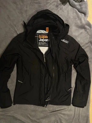 Svart vindjacka från Superdry - Snygg svart vindjacka från Superdry med huva och dragkedja framtill. Jackan har två fickor med dragkedja, ribbad krage och detaljer i vitt på bröstet. Materialet är tunt och vindtätt, perfekt för blåsiga dagar. Storlek M men känns som S.