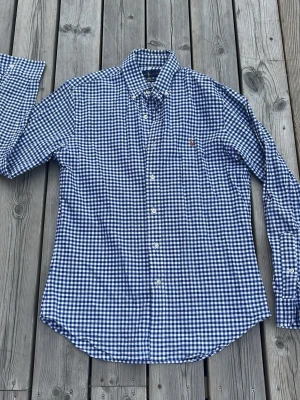 Blårutig skjorta från Polo Ralph Lauren - Klassisk blårutig skjorta från Polo Ralph Lauren med button-down krage och broderad logga på bröstet. Skjortan har långa ärmar och vita knappar framtill. Perfekt för en stilren och avslappnad look.