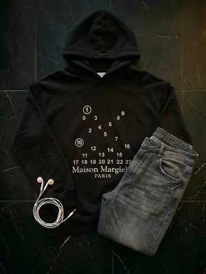 Maison Margiela svart hoodie med tryck - Säljer en svart hoodie från Maison Margiela med ikoniskt siffror-tryck och texten 'Maison Margiela Paris' framtill. Hoodien har huva och ribbade muddar. Perfekt för dig som gillar streetwear och vill sticka ut med en unik design. Stilren hoodie som blir perfekt inför skolstarten. Hoodien är helt oanvänd.