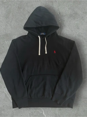  Polo Ralph Lauren Hoodie Svart  - Svart hoodie från Polo Ralph Lauren med klassisk huva och vita dragsnören. På bröstet finns den ikoniska röda Polo-loggan broderad. Hoodien har en stor magficka och ribbade muddar vid ärmar och nederkant. Perfekt för en avslappnad och stilren look. Nyskick! Storlek L
