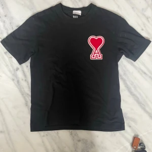 Svart ami Paris T-shirt  - Svart t-shirt i bomull med ett stort rött hjärta och bokstaven A broderat på bröstet. Klassisk passform och korta ärmar, perfekt för en avslappnad och trendig look.