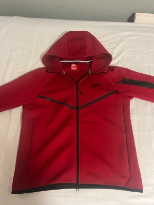 Röd Nike hoodie med dragkedja - Snygg röd hoodie från Nike med hel dragkedja framtill och svart logga på bröstet. Hoodien har huva, svarta detaljer längs sömmarna och en extra ficka med dragkedja på ena ärmen. Perfekt för en sportig och avslappnad stil.