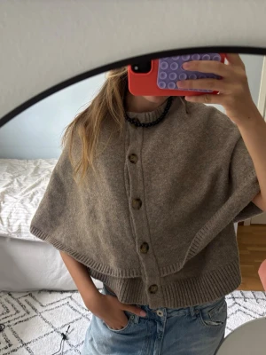 Grå stickad cape med knappar - Säljer en grå stickad cape med ribbade kanter och knappar framtill. Capen har en rund halsringning och är tillverkad i ett mjukt material som känns lyxigt och bekvämt. Perfekt för lager på lager och en snygg detalj till din outfit.