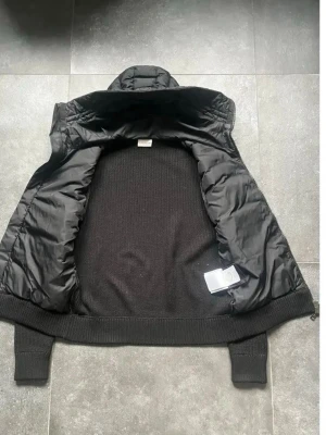 Svart dunväst från Moncler - Säljer en svart dunväst från Moncler med stickad baksida och vadderad framsida. Västen har hög krage, ribbade muddar och dragkedja framtill. Klassisk Moncler-logga på bröstet och cool mix av material som ger en modern look.