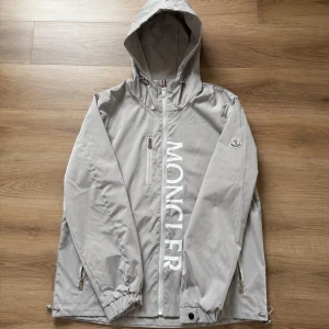Grå vindjacka med huva från Moncler - Snygg grå vindjacka från Moncler med stor vit logga längs dragkedjan framtill. Jackan har huva, dragkedjeförsedda fickor och justerbara muddar. Perfekt för blåsiga dagar och enkel att matcha med olika outfits.