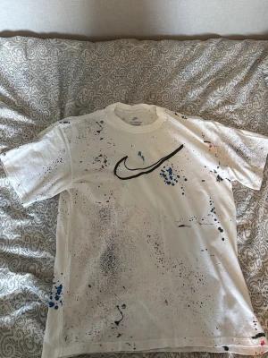 Vit Nike t-shirt  - Säljer en vit t-shirt från Nike med svart logga på bröstet och coola färgstänk i blått, svart och rött över hela tröjan. Baksidan har flera tryck och grafiska detaljer. Perfekt för dig som gillar streetwear och unika plagg.