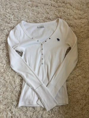 Abercrombie Vit ribbad långärmad topp - Snygg vit ribbad topp med lång ärm och v-ringning. Dekorativa knappar framtill och en liten mörkblå logga broderad på bröstet. Perfekt att styla med jeans eller kjol för en clean look. Från Abercrombie and fitch, storlek XS💙