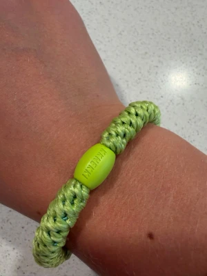 Kknekki hårband - Snyggt armband i limegrönt, flätat av glansigt snöre och med en oval pärldetalj i plast där det står 'RAKELINI'. Perfekt accessoar för att addera färg till din outfit. Passar dig som gillar unika och färgglada smycken.