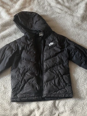 Svart dunjacka med huva från Nike - Snygg svart dunjacka från Nike med huva och dragkedja framtill. Jackan har ett diskret Nike-logga på bröstet och är tillverkad i ett glansigt material. Perfekt för kalla vinterdagar och har en sportig look som funkar till det mesta.