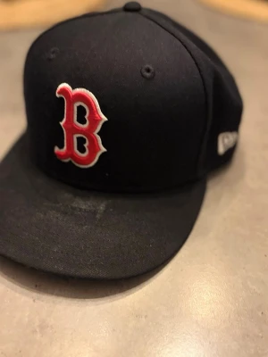 Marinblå Boston Red Sox keps - Snygg svart keps med röd och vit Boston Red Sox-logga broderad framtill. Klassisk modell med böjd skärm och små ventilationshål. Tillverkad i ett slitstarkt tyg, perfekt för dig som gillar sportig stil. Stolek 59,6cm.