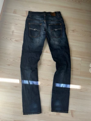 Mörkblå nudie jeans  - Säljer ett par mörkblå jeans med raka ben och slitningar på både fram- och baksida. Jeansen har klassiska fem fickor och kontrastsömmar. Materialet är denim i bomull och passformen är avslappnad men ändå snyggt åtsittande.