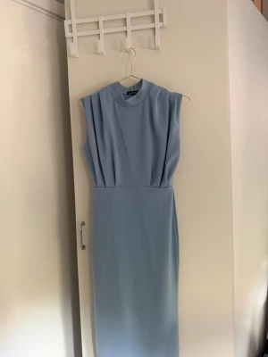 Ljusblå ärmlös långklänning - Säljer en elegant ljusblå långklänning med hög hals och ärmlös design. Klänningen har en rak, smickrande passform och fina veckdetaljer framtill upptill. Perfekt för dig som gillar stilrena och minimalistiska plagg.