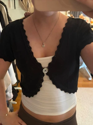 Svart kortärmad kofta med knapp - Söt svart kortärmad kofta med scallop-kant och en stor vit knapp framtill. Modellen är croppad och har en djup v-ringning, perfekt att bära över linne eller topp. Skriv pm vid funderingar!☺️