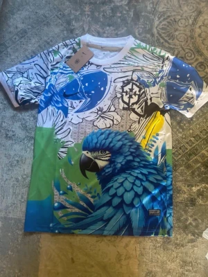 Färgstark t-shirt med papegojtryck - Unik t-shirt med stor blå papegoja framtill och färgglada detaljer i blått, grönt, vitt och gult. T-shirten har korta ärmar, rund hals och ett coolt grafiskt mönster med blommor och blad. Perfekt för dig som vill sticka ut med din stil.