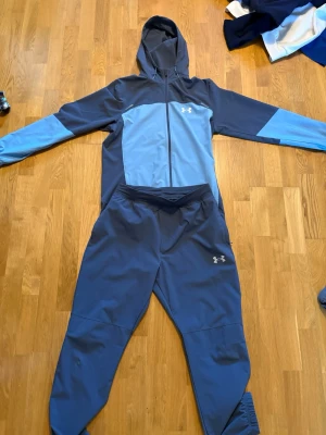 Blå träningsset från Under Armour - Snyggt träningsset från Under Armour i olika blå nyanser. Setet består av en hoodie med dragkedja och matchande byxor. Materialet är troligtvis polyester, perfekt för träning eller chill. Under Armour-loggan syns på både jackan och byxorna.