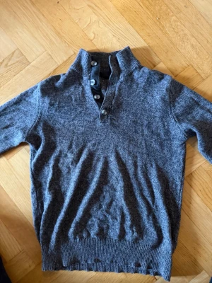 Mörkgrå stickad tröja med knappar - Mysig mörkgrå stickad tröja med hög krage och fyra knappar framtill. Tröjan har långa ärmar och ribbade muddar vid ärmslut och nederkant. Perfekt för lager på lager och en avslappnad stil.