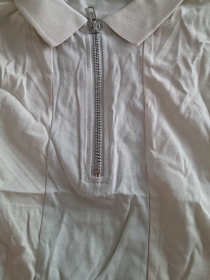 Begie polotopp med half zip - Snygg vit polotopp med krage och half zip-dragkedja framtill. Tillverkad i mjukt bomullsmaterial som känns skönt mot huden. Perfekt till jeans eller kjol för en clean och fräsch look.