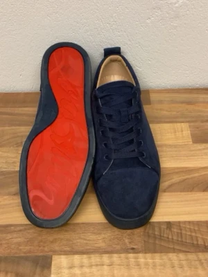 Mörkblå mocka sneakers med röd sula - Snygga mörkblå sneakers i mocka med klassisk röd sula och ton-i-ton snörning. Skorna har broderad logga bak och en stilren, låg profil. Perfekta för dig som gillar exklusiv och trendig streetstyle.