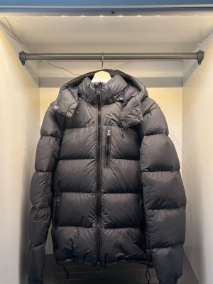 Mörkgrå dunjacka från Polo Ralph Lauren - Snygg mörkgrå dunjacka från Polo Ralph Lauren med avtagbar huva, dragkedja framtill och flera praktiska fickor. Jackan har broderad logga på bröstet och är perfekt för kalla vinterdagar. Tjockt och puffigt material som håller dig varm. Använd 1 vinter