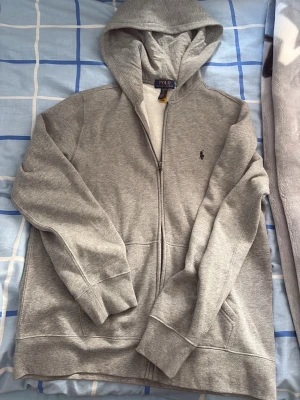 Grå hoodie från Polo Ralph Lauren - Snygg grå hoodie från Polo Ralph Lauren med dragkedja framtill och klassisk huva. Tröjan har två fickor och den ikoniska lilla loggan broderad på bröstet. Tillverkad i mjukt bomullsmaterial som är perfekt för chill dagar.