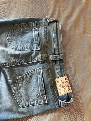 True Religion Becca Bootcut Jeans - Snygga blå jeans från True Religion, modell Becca Mid Rise Bootcut. Klassisk bootcut-passform med tydliga sömmar och ikoniska bakfickor med broderad detalj. Perfekta för dig som gillar en avslappnad men trendig look.