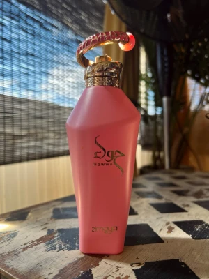 Zimaya Hawwa Rosa; 100ml - Unik parfymflaska i matt rosa med gulddetaljer och ett dekorativt handtag i guld och rosa. Flaskan har en modern, kantig form och texten 'Hawwa' samt 'Zimaya' i guld på framsidan. Perfekt för dig som gillar snygg design på parfymhyllan. Bara testad, som ny.