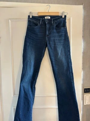 Blå bootcut jeans från Wrangler - Snygga blå bootcut jeans från Wrangler med klassisk femficksmodell och medelhög midja. Jeansen har en mörk tvätt och midjemåttet tvärs över är 33cm. Perfekta för dig som gillar en retroinspirerad look med utsvängda ben.